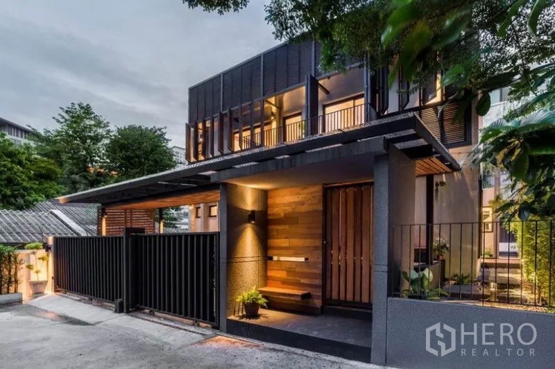 Townhouse for rent in Khlong Toei, Bangkok - Modern two-storey townhouse exterior at dusk with wood-and-metal facade, secure gate and warm lighting.