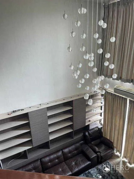 Townhouse for rent in Khlong Toei, Bangkok - Double-height living room with leather sectional sofa, wall shelves, and cascading chandelier viewed from above.