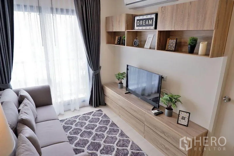 Condominium for rent in Huai Khwang, Bangkok - Modern living room with sofa, TV wall unit, and balcony door with sheer curtains.