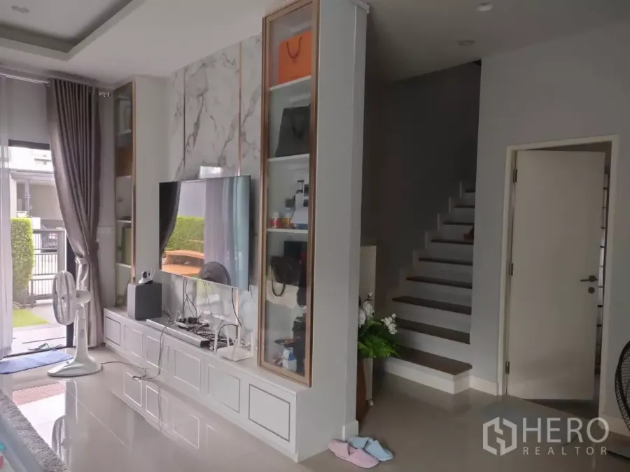 Detached House for rent or sale in Lam Luk Ka, Pathum Thani - Modern living room with marble TV wall, built‑in shelves, and staircase beside a sliding glass door.