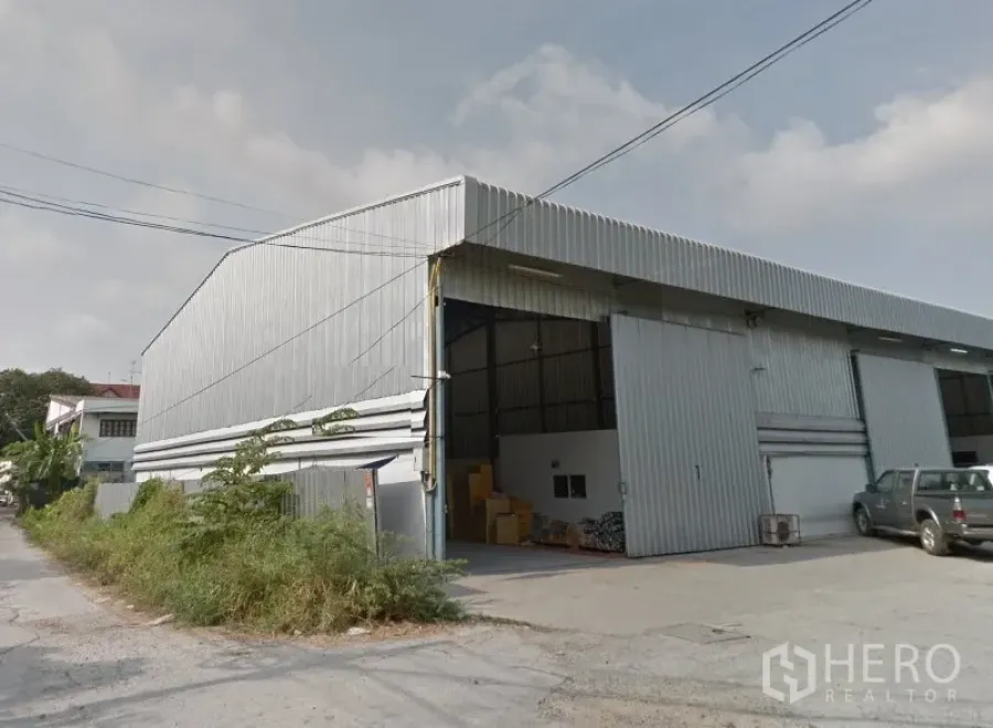 Warehouse for rent in Pak Kret, Nonthaburi - Exterior of a corrugated metal warehouse in Pak Kret with high gable roof, wide sliding doors, and a concrete loading area on a quiet side street.