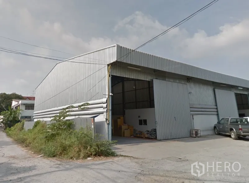 Warehouse for rent in Pak Kret, Nonthaburi - Exterior of a corrugated metal warehouse in Pak Kret with high gable roof, wide sliding doors, and a concrete loading area on a quiet side street.
