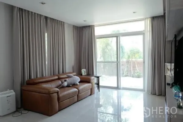 บ้าน ขายในPak Kret, Nonthaburi - ห้องรับแขกสว่างด้วยประตูกระจกบานเลื่อน ผ้าม่านสีเทา และโซฟาหนังสีน้ำตาล
