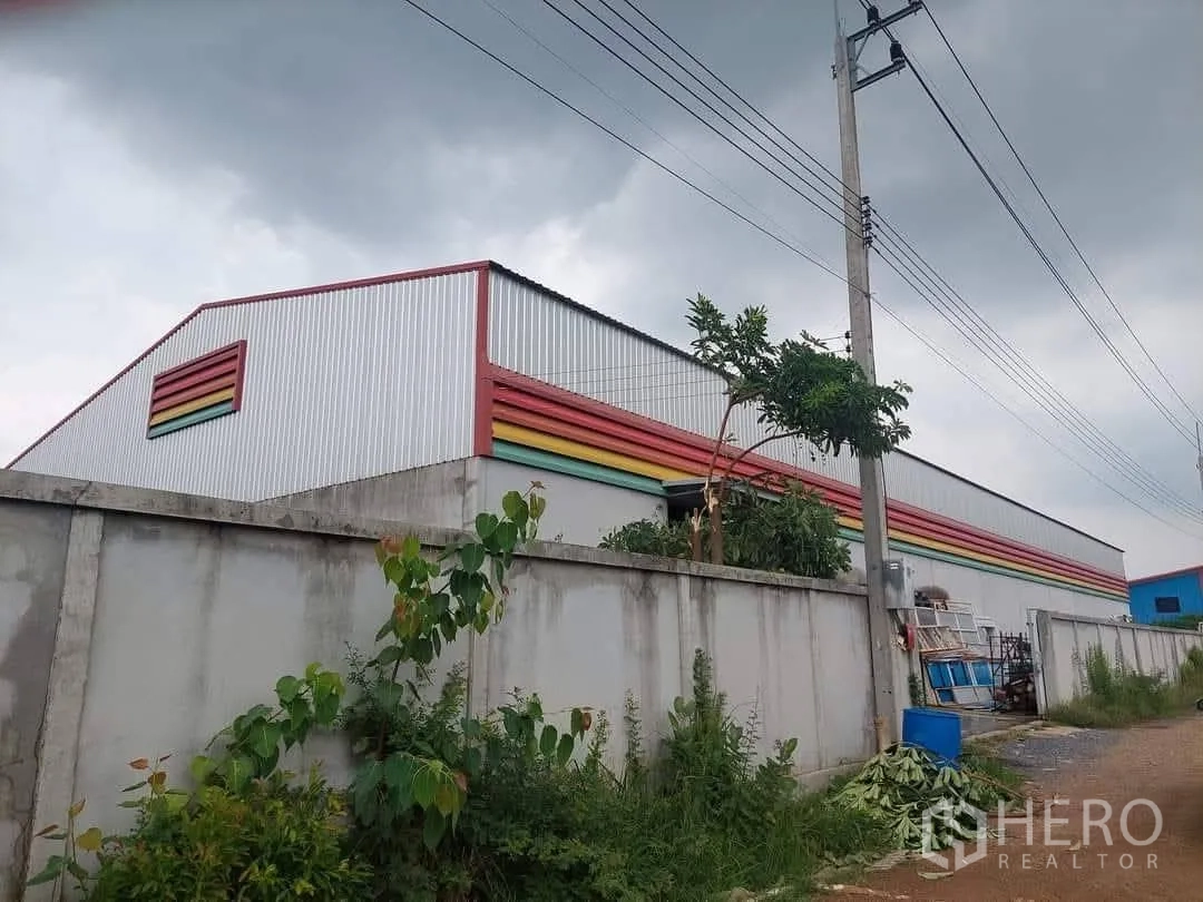 Warehouse for sale in Lam Luk Ka, Pathum Thani - Angled exterior view of the warehouse’s high gable roof, perimeter wall, and roadside vegetation.