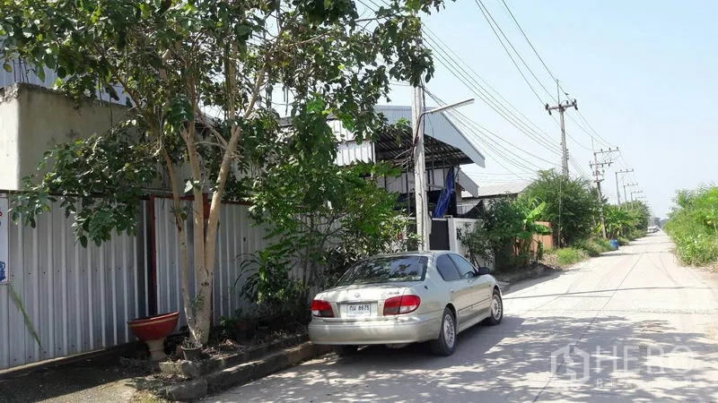 Warehouse for sale in Pathum Thani - Quiet access road with trees, power lines and on‑street parking outside the warehouse.