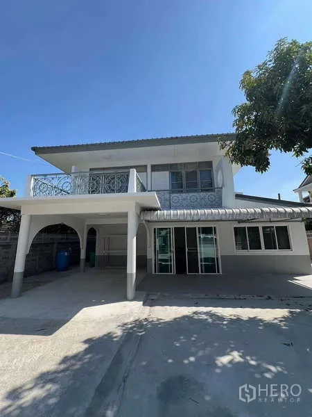 Detached House for rent in Bang Na, Bangkok - Two-storey detached house exterior with carport, balcony railing and a wide paved driveway under a blue sky.