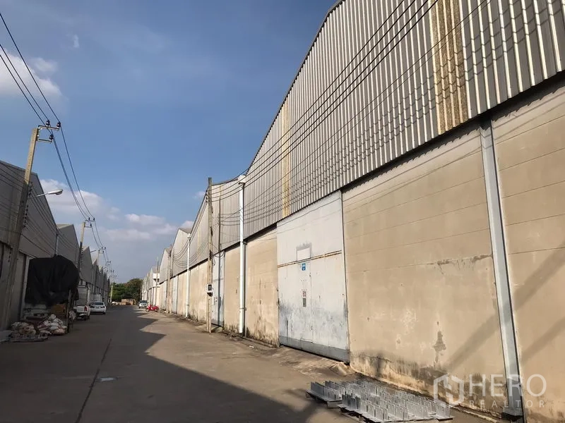 Warehouse for rent in Bang Phli, Samut Prakan - Side view of warehouse blocks with closed metal doors along a wide access road and overhead wiring.
