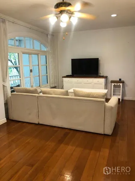 Townhouse for rent in Mueang Samut Prakan, Samut Prakan - Living room with large sofa facing TV console, ceiling fan, wood floors and French doors opening to a balcony.