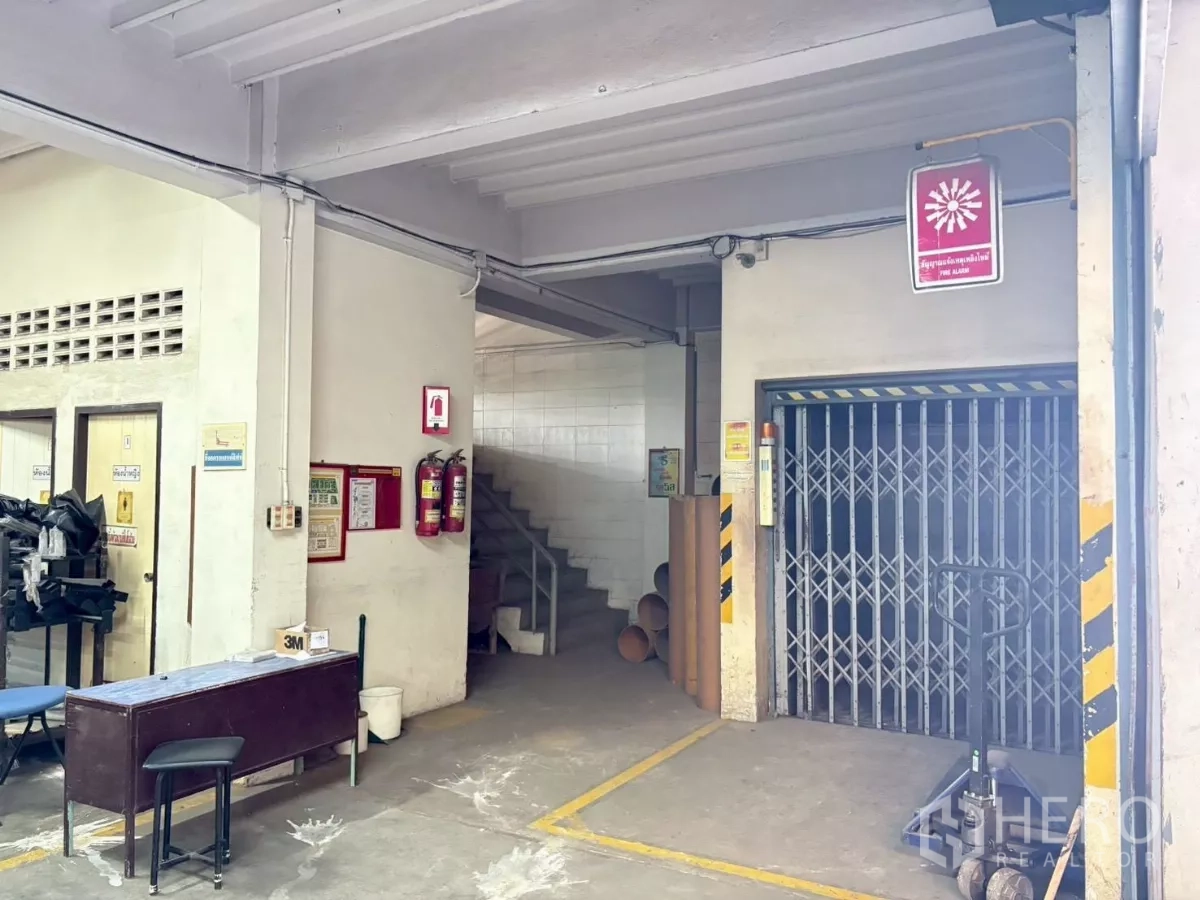 Factory for rent or sale in Bang Khun Thian, Bangkok - Ground-floor zone with folding-gate goods lift, staircase and safety signage.