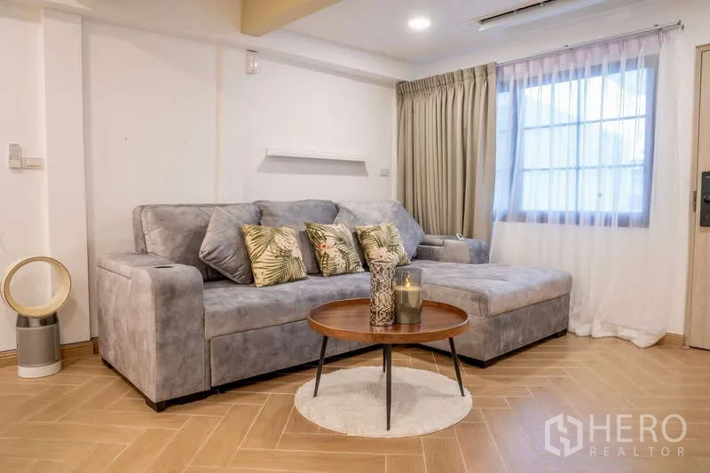 Townhouse for rent in Khlong Toei, Bangkok - Living room with grey L-shaped sofa, sheer curtains and a round coffee table.