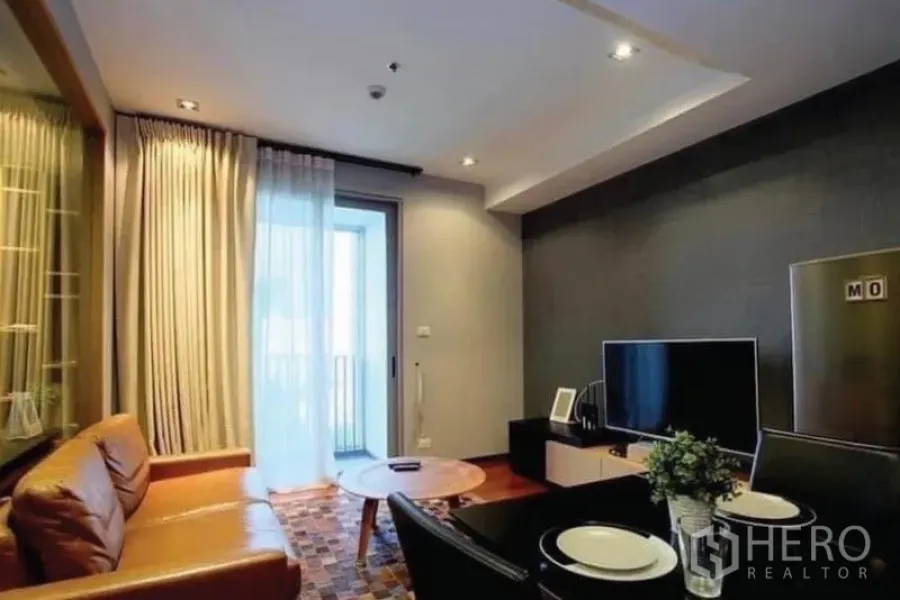 Condominium for sale in Khlong Toei, Bangkok - Cozy living area with brown leather sofa, TV console, and a balcony door bringing in natural light.