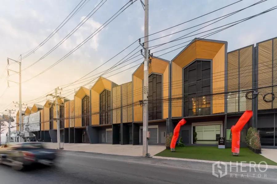 Warehouse for rent in Mueang Samut Sakhon, Samut Sakhon - Row of modern loft-style warehouses with gabled facades facing a main road, wide driveways and red air dancers at the entrance at dusk.