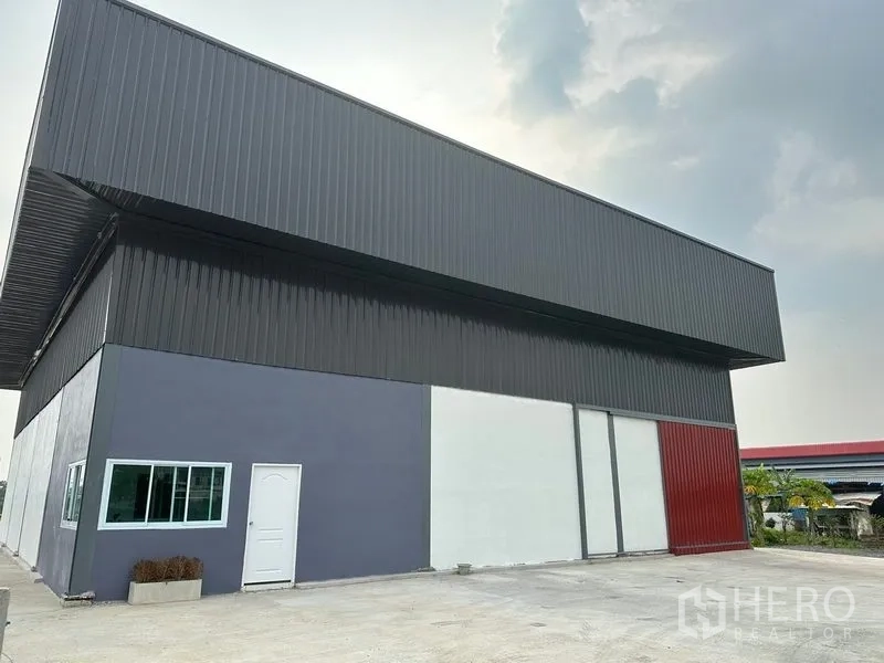Warehouse for rent in Bang Bua Thong, Nonthaburi - Angled view of a large industrial warehouse with deep metal roof overhang, side entry door, windows and a fenced driveway.