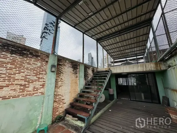 Shophouse for rent in Bang Rak, Bangkok - Rooftop terrace with metal stairs and a covered deck, city skyline visible beyond the brick wall.
