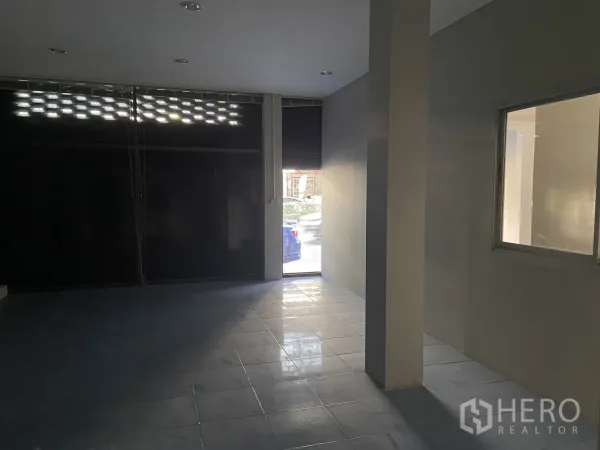 Shophouse for rent in Bang Na, Bangkok - Front retail space with tiled floor and a partially open roller door allowing daylight.