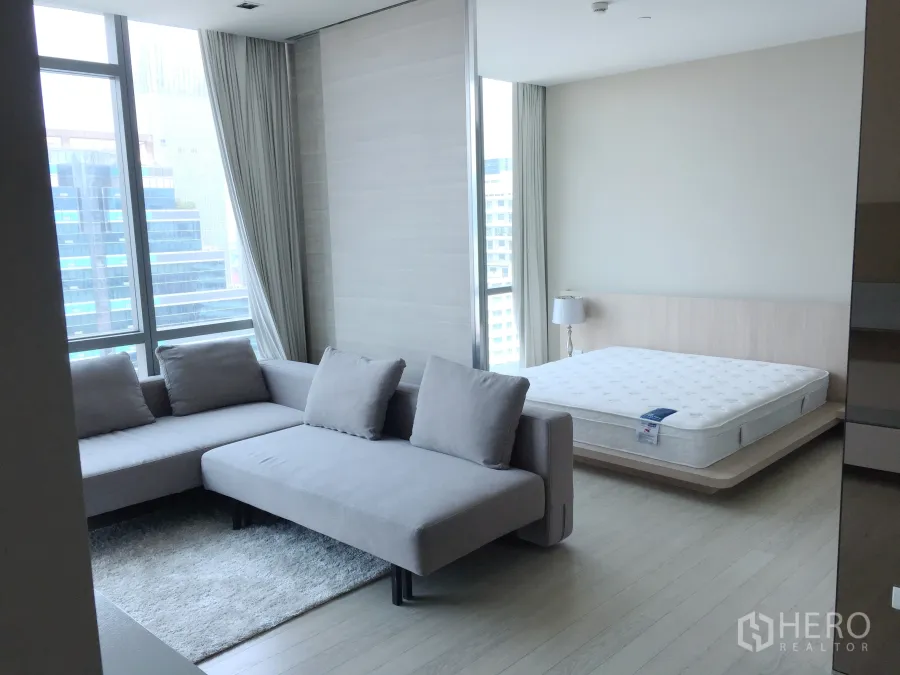 Condominium for rent or sale in Watthana, Bangkok - Bright open-plan living and sleeping area with L-shaped sofa, platform bed and floor-to-ceiling windows with city view.