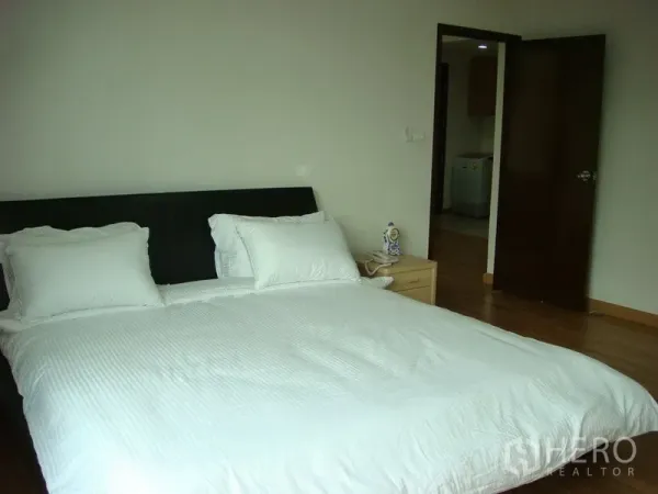 Apartment for rent in Khlong Toei, Bangkok - Master bedroom with white bedding, wood flooring, bedside table, and an open doorway in a Bangkok apartment.