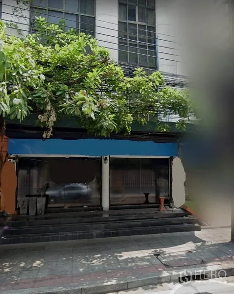 Shophouse for rent in Watthana, Bangkok - Street-facing shophouse facade with wide glass storefront, entry steps and leafy tree canopy in Watthana.