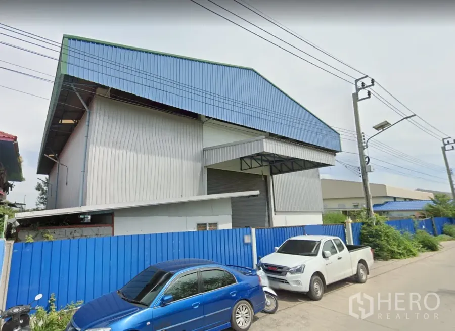 Warehouse for rent in Pak Kret, Nonthaburi - Street view of a metal-clad warehouse with a blue roof, wide roller shutter, covered loading bay, and gated frontage in Pak Kret, Nonthaburi.