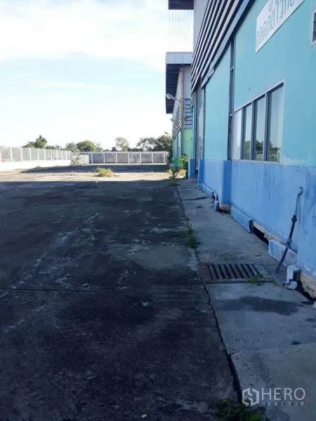 Factory for rent in Samut Prakan - Side exterior of blue industrial warehouse with wide concrete yard and drainage in Bang Sao Thong, Samut Prakan.