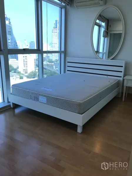 Condominium for rent in Khlong Toei, Bangkok - Bright bedroom with floor-to-ceiling windows, city view, white bed and round mirror.