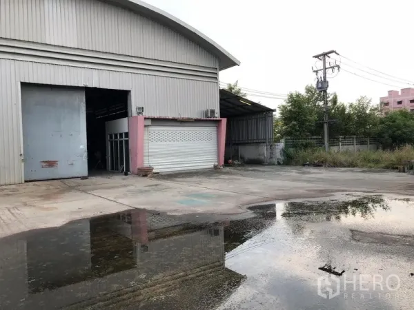 Warehouse for rent or sale in Lam Luk Ka, Pathum Thani - Steel-clad warehouse exterior with large roller shutter and wide concrete yard with drainage, fencing and power lines in a green setting.