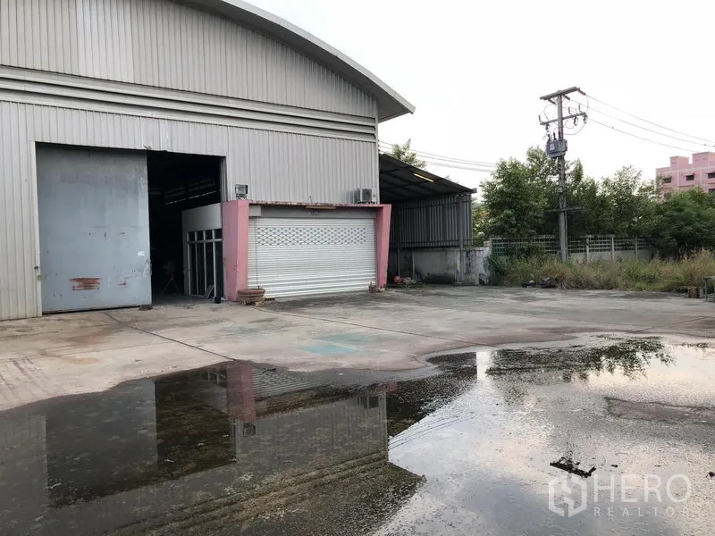 Warehouse for rent or sale in Lam Luk Ka, Pathum Thani - Steel-clad warehouse exterior with large roller shutter and wide concrete yard with drainage, fencing and power lines in a green setting.