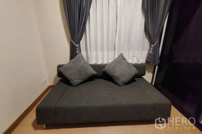 Townhouse for rent in Thanyaburi, Pathum Thani - Small lounge corner with dark sofa and full-length curtained window.