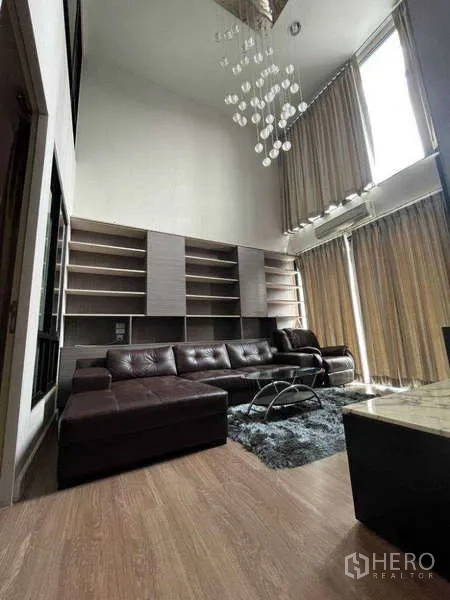 Townhouse for rent in Khlong Toei, Bangkok - Living area with high ceiling, brown sectional sofa, bookshelves, and floor-to-ceiling drapes.