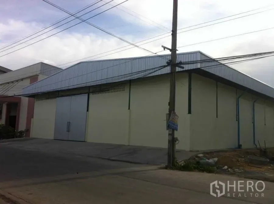 Warehouse for rent in Bang Phli, Samut Prakan - Angled view of the warehouse exterior showing wide driveway, utility lines, and corrugated roof.