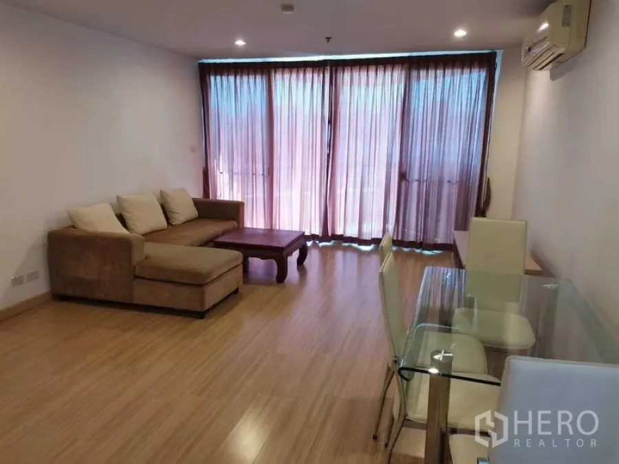 Condominium for rent in Khlong Toei, Bangkok - Bright living–dining area with L‑shaped sofa, glass table, wood floors and full‑height curtained windows.