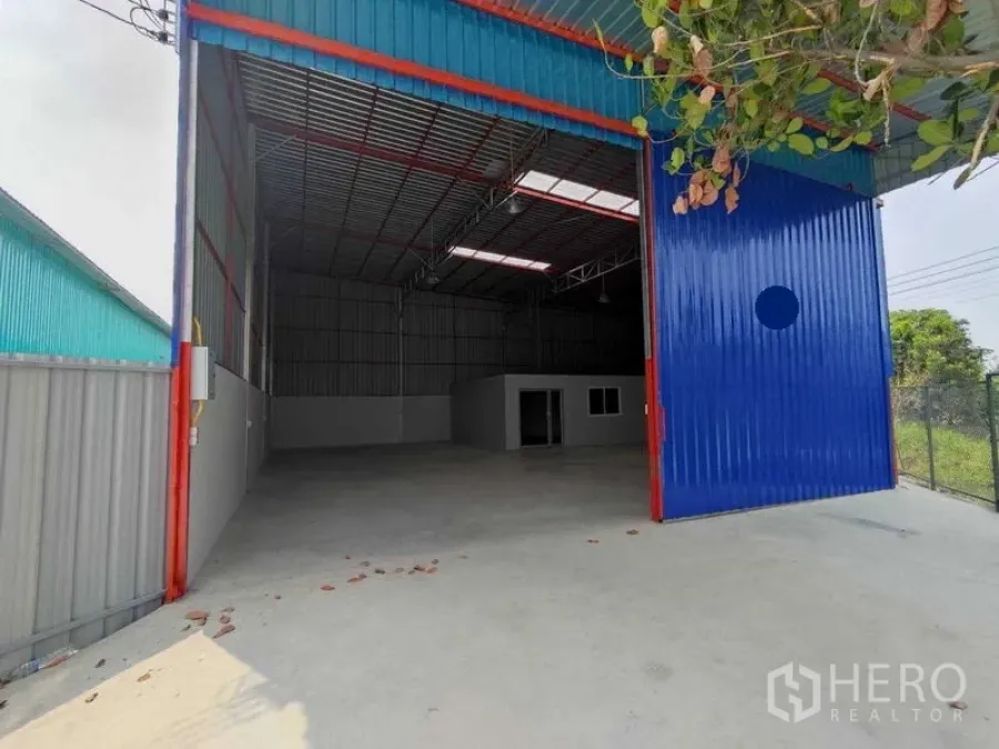 Warehouse for rent in Pak Kret, Nonthaburi - High-clearance warehouse with large blue sliding door, concrete apron, and an internal office visible.