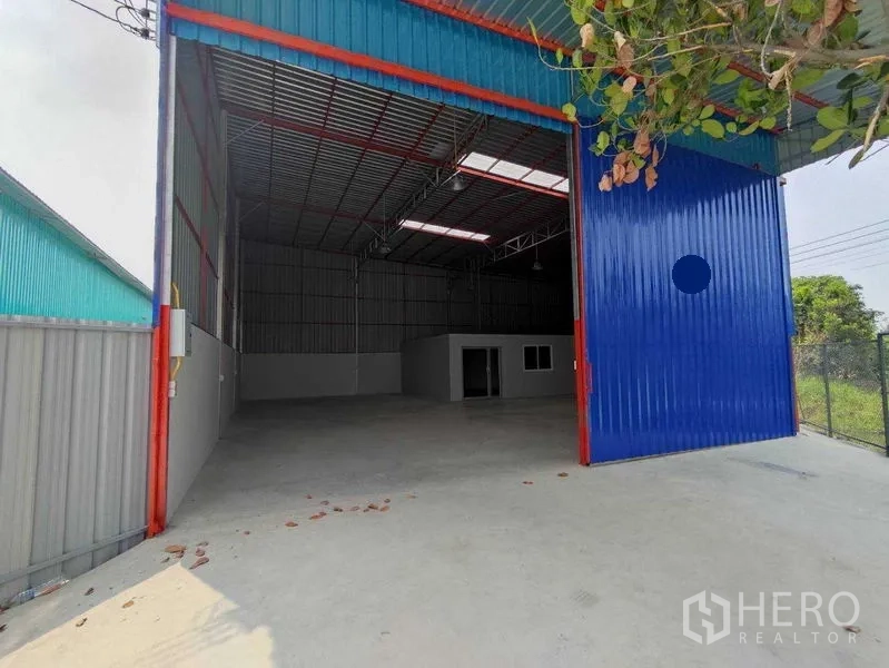 Warehouse for rent in Pak Kret, Nonthaburi - High-clearance warehouse with large blue sliding door, concrete apron, and an internal office visible.