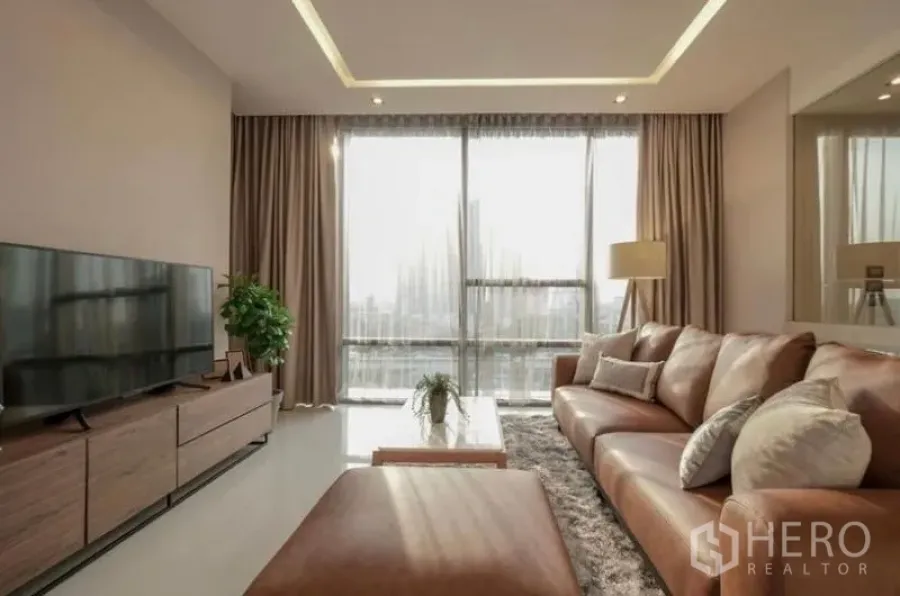 Condominium for rent in Sathon, Bangkok - Sunlit living room with floor-to-ceiling windows, leather sectional sofa, TV console and city view.