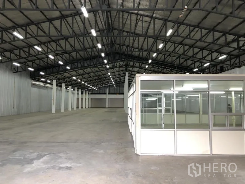 Warehouse for rent in Nonthaburi - Warehouse hall with a glass office block in the foreground, even lighting and a smooth concrete slab.