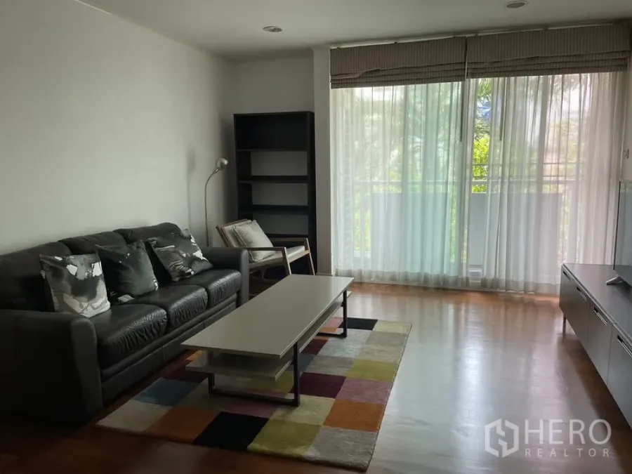 Condominium for rent in Pathum Wan, Bangkok - Bright living room with black leather sofa, coffee table, colorful rug and floor‑to‑ceiling windows opening to greenery in Lumphini, Pathum Wan, Bangkok.
