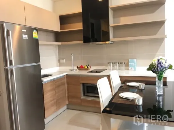 Condominium for rent in Khlong Toei, Bangkok - L‑shaped kitchen with wood cabinets, stainless fridge, cooktop, open shelves and a two‑seat dining counter.