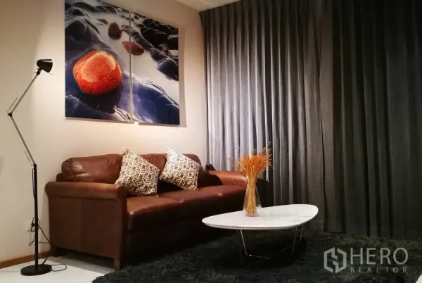 Condominium for rent or sale in Khlong Toei, Bangkok - Contemporary living room with a brown leather sofa, bold rock artwork, floor lamp and oval marble coffee table against dark drapes.