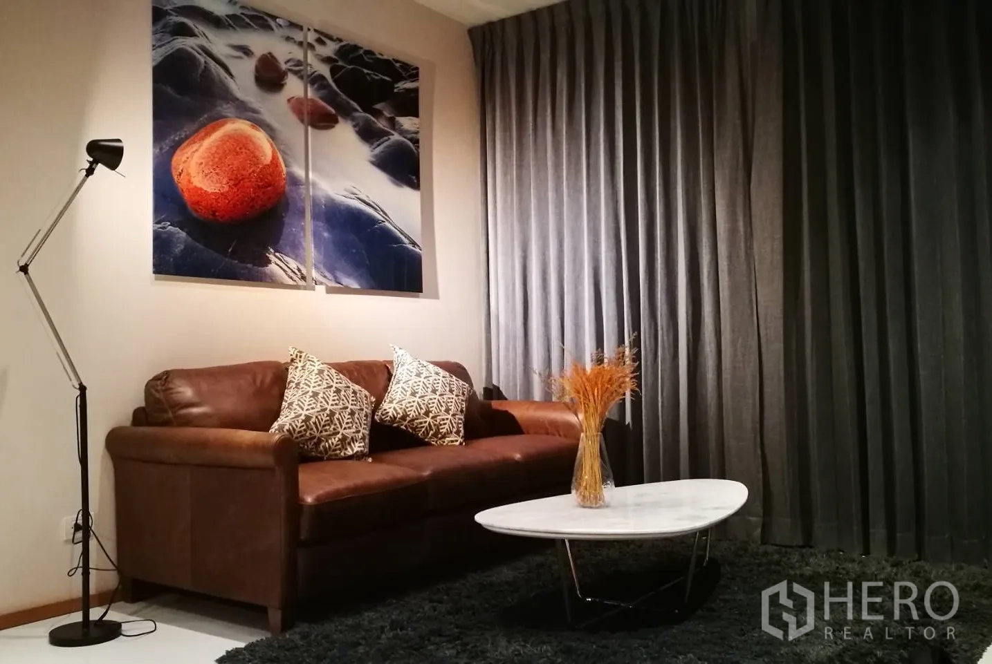 Condominium for rent or sale in Khlong Toei, Bangkok - Contemporary living room with a brown leather sofa, bold rock artwork, floor lamp and oval marble coffee table against dark drapes.