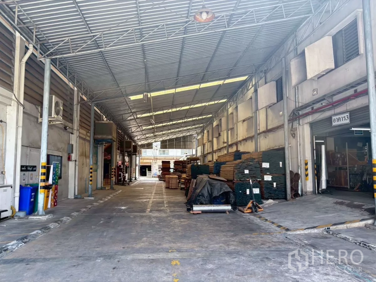 Factory for rent or sale in Bang Khun Thian, Bangkok - Covered loading lane between buildings with stored materials and building signage.
