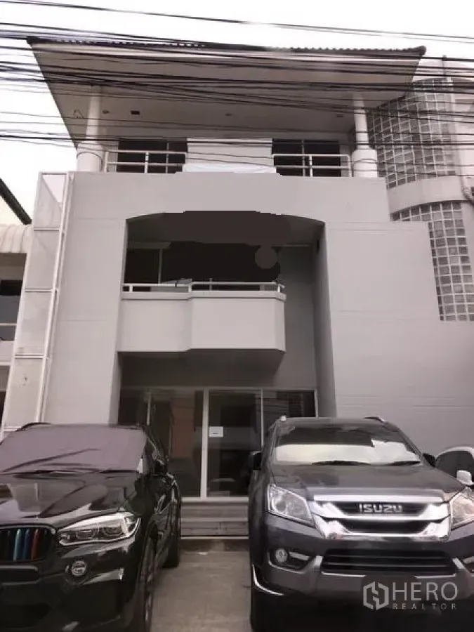 Home Office for rent in Huai Khwang, Bangkok - Three-storey home office exterior with a small front balcony, glass-block corner wall, and parking space with cars in front.