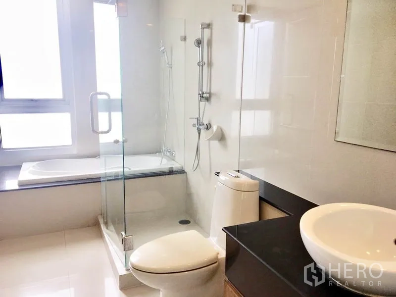 Condominium for rent in Khlong Toei, Bangkok - Modern bathroom with glass shower, bathtub, window and countertop basin.