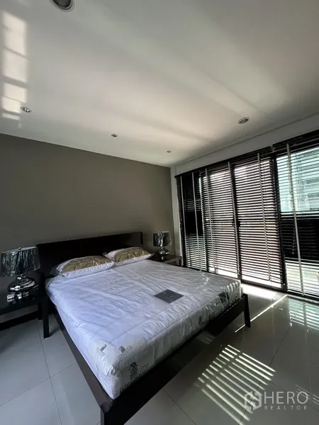 Condominium for rent in Khlong Toei, Bangkok - Second bedroom with king bed, full-height glazing, and balcony access with blinds.