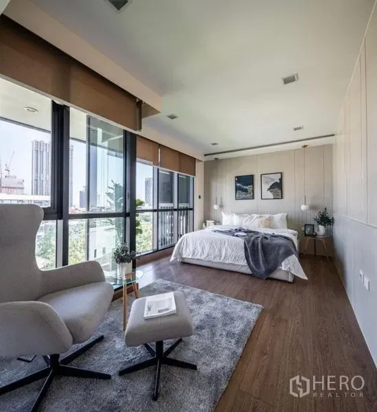 Condominium for rent or sale in Phra Khanong, Bangkok - Spacious master bedroom with panoramic corner windows, lounge chair and ottoman on a soft rug, overlooking the city.