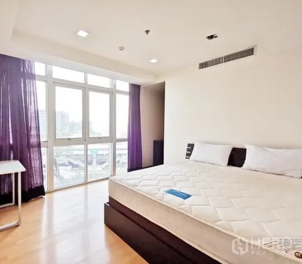 Condominium for rent in Khlong Toei, Bangkok - Corner bedroom with city views through large windows, wood flooring and a king‑size bed.