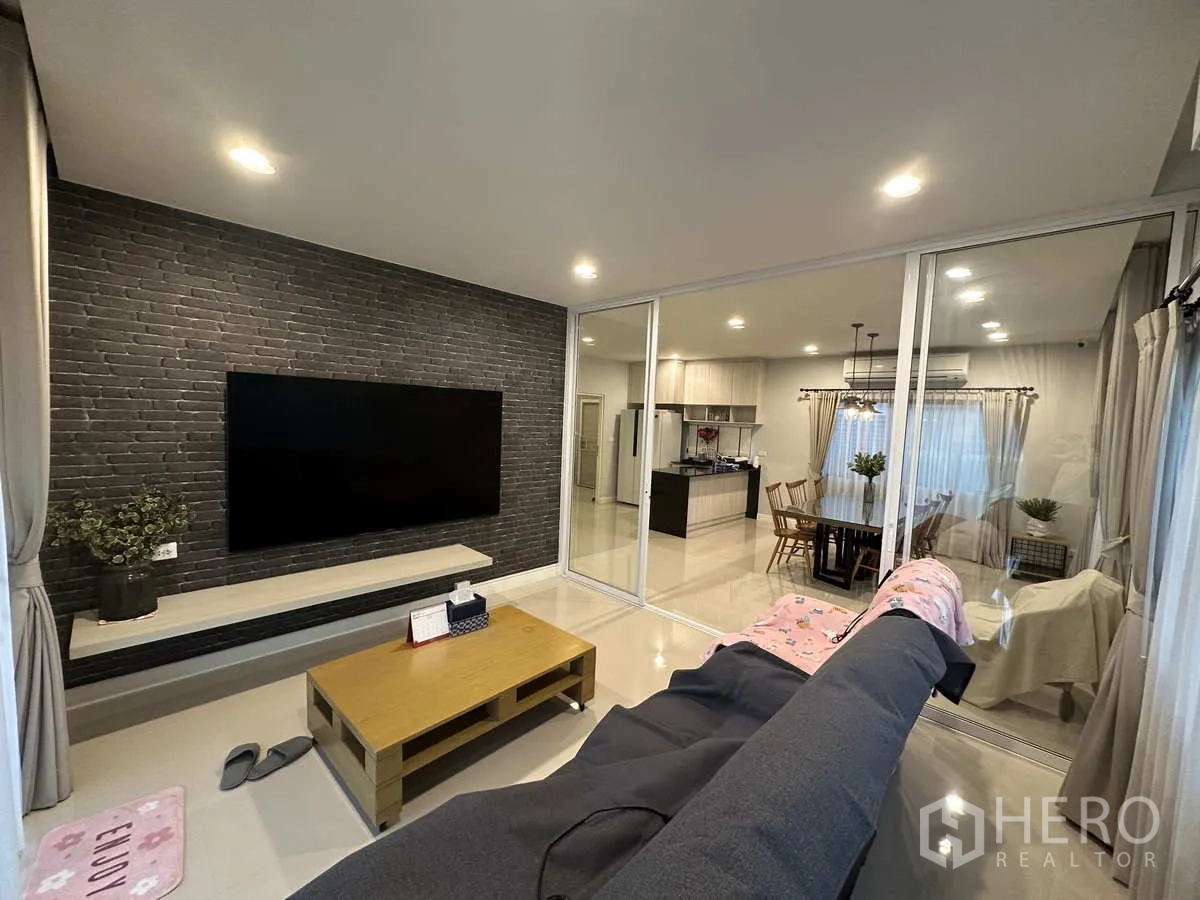 Detached House for rent or sale in Lam Luk Ka, Pathum Thani - Cozy living room with brick accent wall, large TV and glass partition doors.