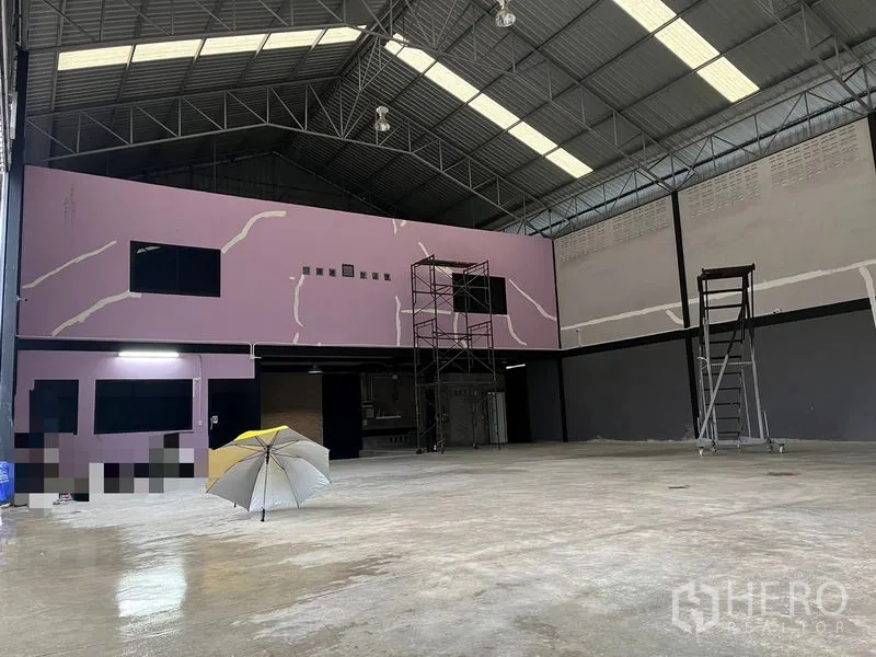 Warehouse for rent in Lat Krabang, Bangkok - Wide view of high-bay warehouse with skylight roof panels, purple mezzanine-style office block, and scaffolding on the floor.