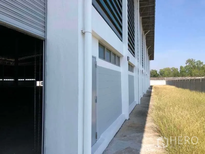 Factory for rent or sale in Phra Nakhon Si Ayutthaya - Side facade with ventilation louvers, a roller shutter door and grass setback along the fence line.