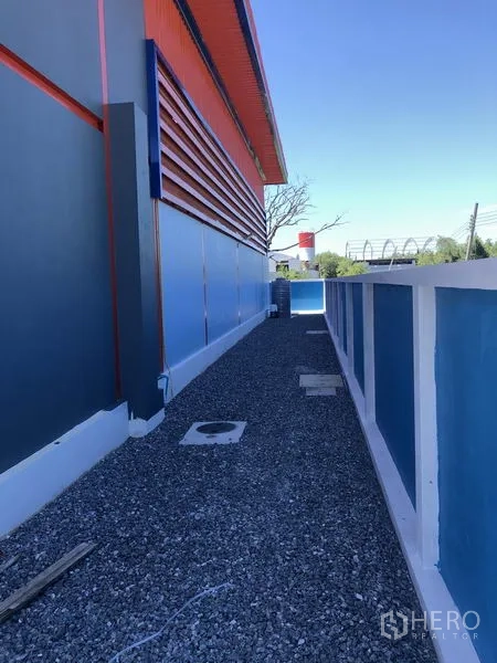 Warehouse for rent or sale in Sai Noi, Nonthaburi - Side gravel walkway along the warehouse exterior with blue boundary wall.