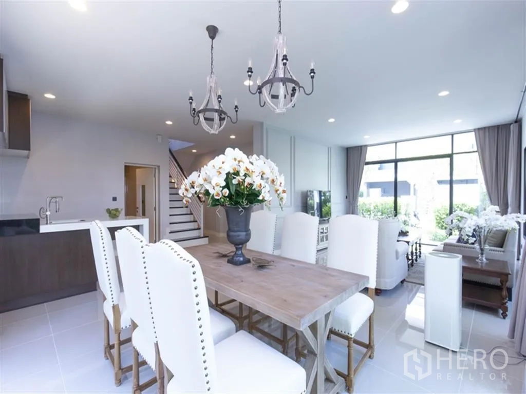 Detached House for rent in Bang Kapi, Bangkok - Dining space with wooden table, white upholstered chairs, and pendant lights beside the kitchen and stairs.
